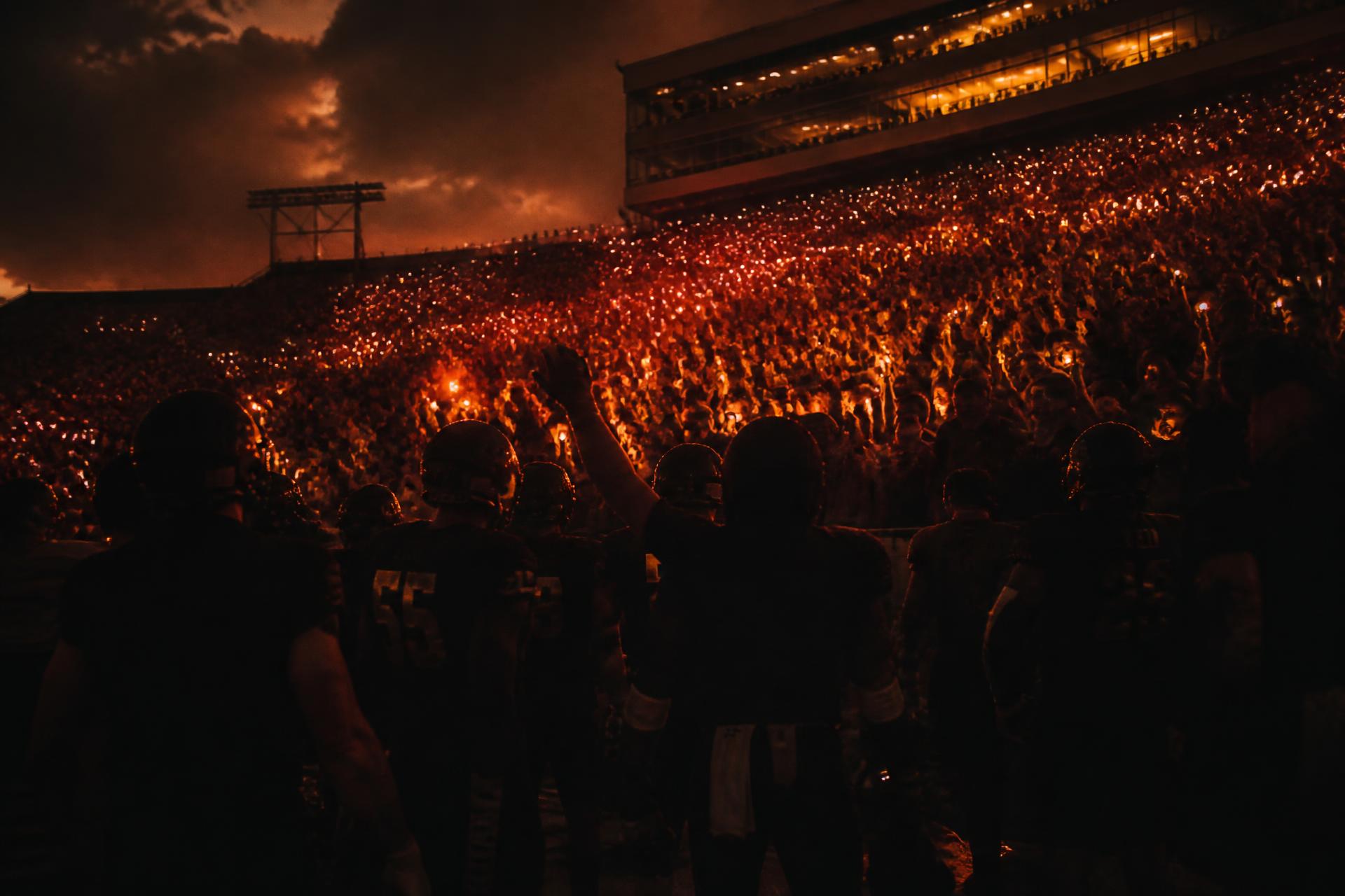 A packed college stadium at dusk, lit by thousands of small amber lights from the crowd